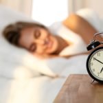 Beautiful young woman sleeping and smiling while lying in bed comfortably and blissfully on the background of alarm clock is going to ring. Sunbeam dawn  on her face.