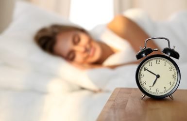 Beautiful young woman sleeping and smiling while lying in bed comfortably and blissfully on the background of alarm clock is going to ring. Sunbeam dawn  on her face.